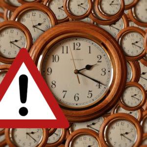 Na pierwszym planie tradycyjny zegar w drewnianej oprawie tarczy, w tle ten sam zegar mniejszy i powielony wielokrotnie W lewym rogu w trójkącie z czerwoną ramką czarny wykrzyknik