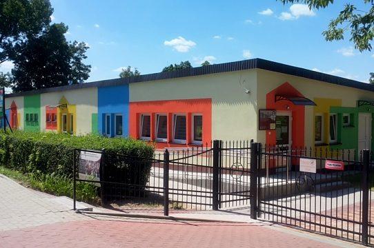 Zza ogrodzenia i żywopłotu widoczny niski biały parterowy budynek pomalowany w kolorowe prostokąty