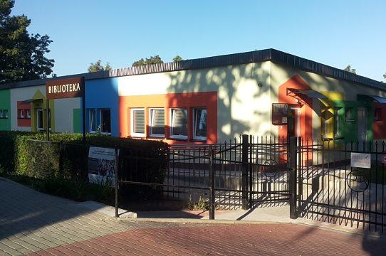 Zza ogrodzenia i żywopłotu widoczny niski biały parterowy budynek pomalowany w kolorowe prostokąty