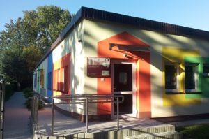 Obramowane szerokim pomarańczowym pasem wejście z logo biblioteki do niskiego parterowego budynku Z lewej i prawej strony kolorowo obramowane okna Z boku podjazd dla wózków