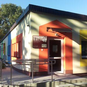 Obramowane szerokim pomarańczowym pasem wejście z logo biblioteki do niskiego parterowego budynku Z lewej i prawej strony kolorowo obramowane okna Z boku podjazd dla wózków