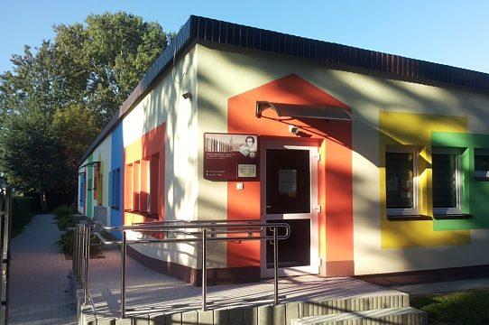 Obramowane szerokim pomarańczowym pasem wejście z logo biblioteki do niskiego parterowego budynku Z lewej i prawej strony kolorowo obramowane okna Z boku podjazd dla wózków