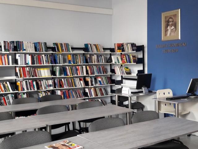 Widok na szare stoliki czytelni. Z prawej strony stanowiska komputerowe, nad nimi, na niebieskim tle ściany portret Juliusza Słowackiego. W tle regały.