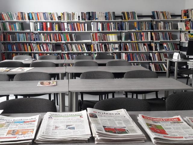 Na pierwszym planie czasopisma bieżące, w tle szare stoliki i krzesła czytelni, wzdłuż ściany regały z książkami.