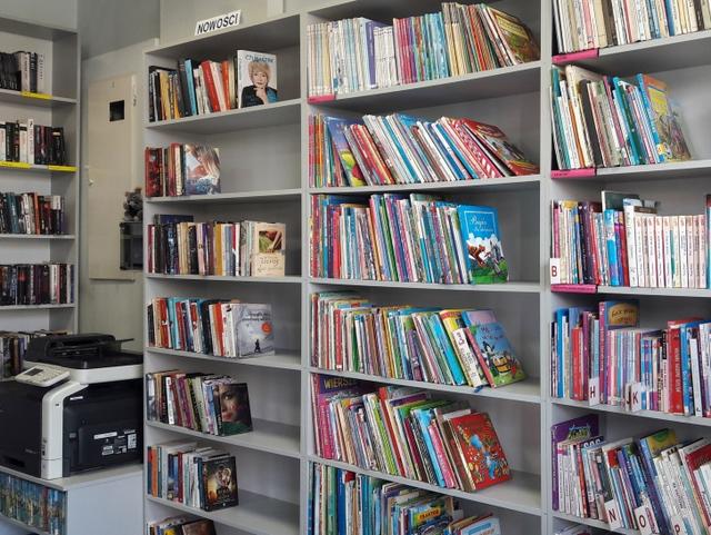 Na pierwszym planie regały z książkami dla dzieci oraz nowościami. W tle urządzenie wielofunkcyjne.