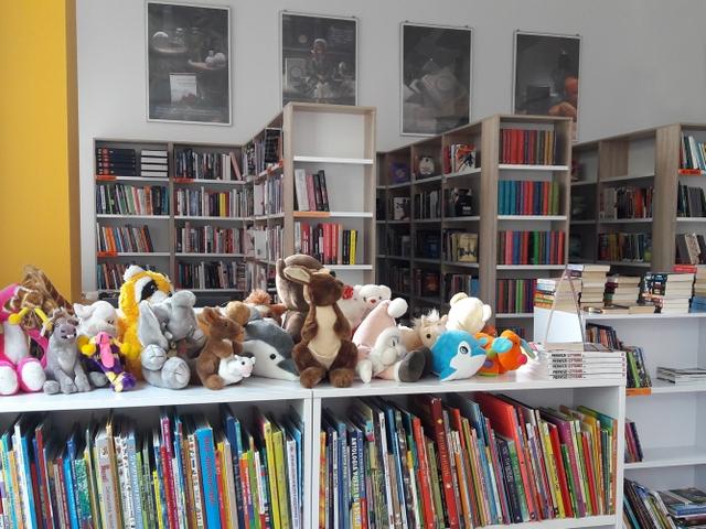Na pierwszym planie niski regał z książeczkami dla dzieci. Nad nim cała półka pluszaków. W tle regały i stylizowane zdjęcia związane z literaturą