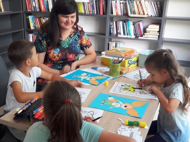 Przy stoliku czytelni bibliotekarka i trójka dzieci z kolorowankami.
