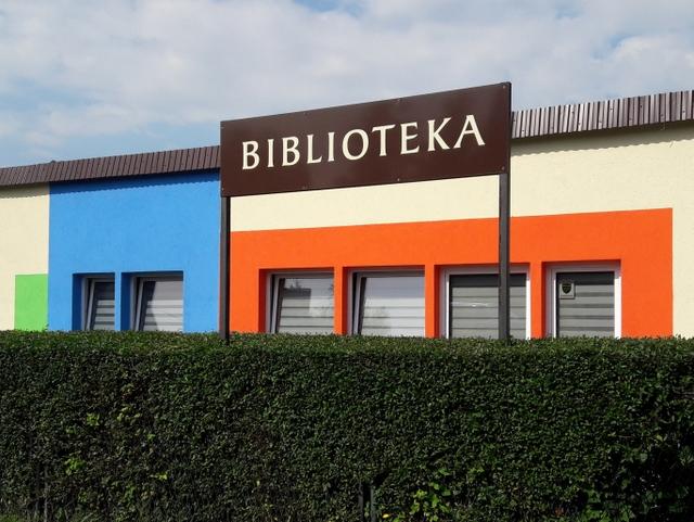 Zza żywopłotu widoczny kolorowy budynek filii z napisem "Biblioteka"