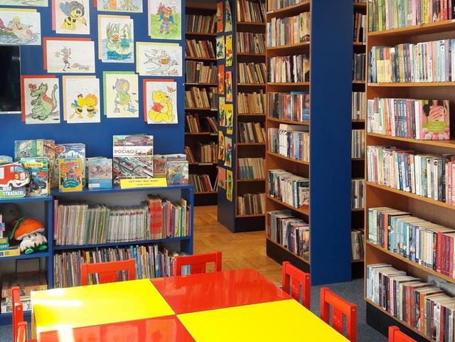 Widok na czytelnię. Na pierwszym planie kolorowe stoliki z krzesełkami dla dzieci. W tle wystawka kolorowanek. Z lewej strony regały.