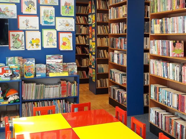Widok na czytelnię. Na pierwszym planie kolorowe stoliki z krzesełkami dla dzieci. W tle wystawka kolorowanek. Z lewej strony regały.