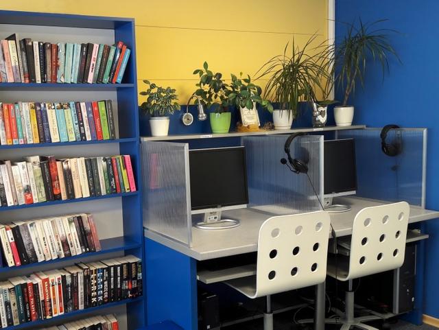 Stanowiska komputerowe dla czytelników. Nad monitorami półka z kwiatami. Obok regał z nowościami książkowymi.