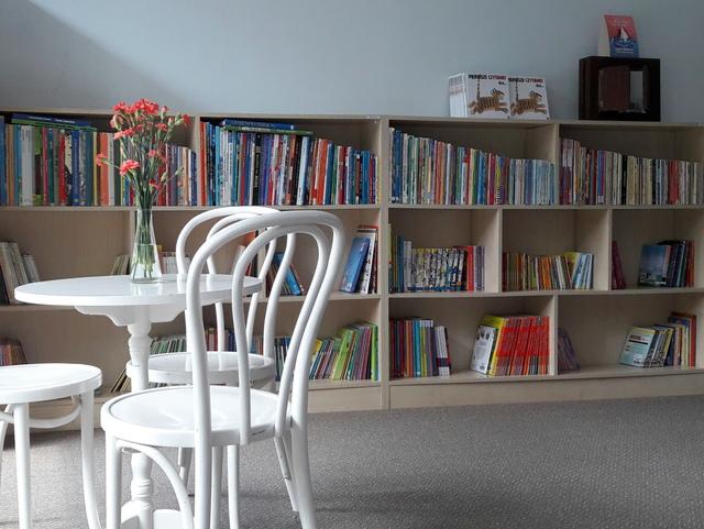 Na pierwszym planie biały okrągły stolik z trzema krzesłami, na nim bukiet kwiatów. W tle niskie regały z książkami dla dzieci.