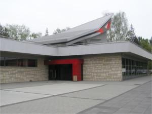 Obramowane na czerwono wejście do budynku Widoczny fragment ukośnego dachu Całość w szaro- beżowej kolorystyce