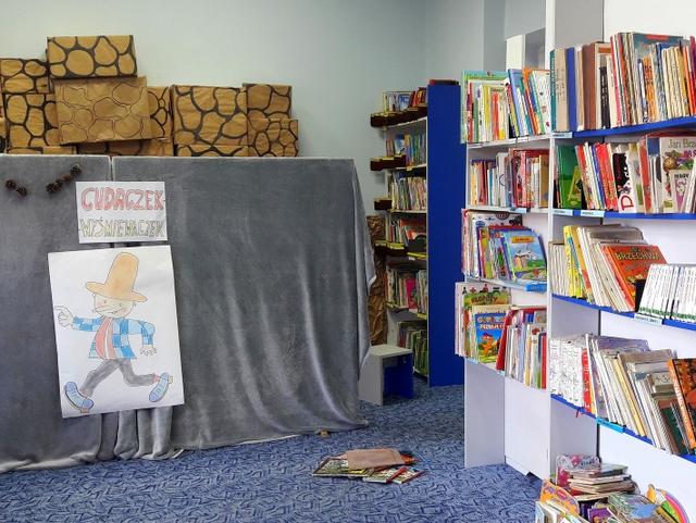 Fragment półek z książkami, z lewej strony przysłonięty szarą tkaniną teatrzyk do przedstawień lalowych.