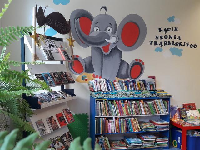Na ścianie namalowany duży, siedzący słoń. Obok napis Kącik Słonia Trąbalskiego. W dole zdjęcia niski regał z książkami dla dzieci.