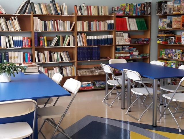 Widok na czytelnię: niebieskie stoliki z białymi krzesłami. W tle regały z książkami i grami planszowymi.