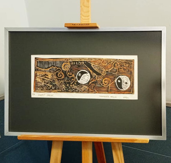 Na sztalugach obraz w cienkiej szarej ramie i szerokim czarnym passepartout. Linoryt. Obraz utrzymany w tonacjach brązów i beżów, ze stylizowanymi sylwetkami ptaków. W prawym dolnym rogu symbol yin i yang.