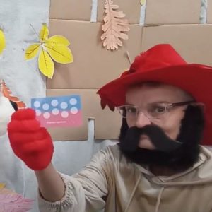 Postać w czerwonym kapeluszu i czerwonych rękawiczkach z doklejoną czarną brodą i wąsami pokazuje kartę z kółeczkami.