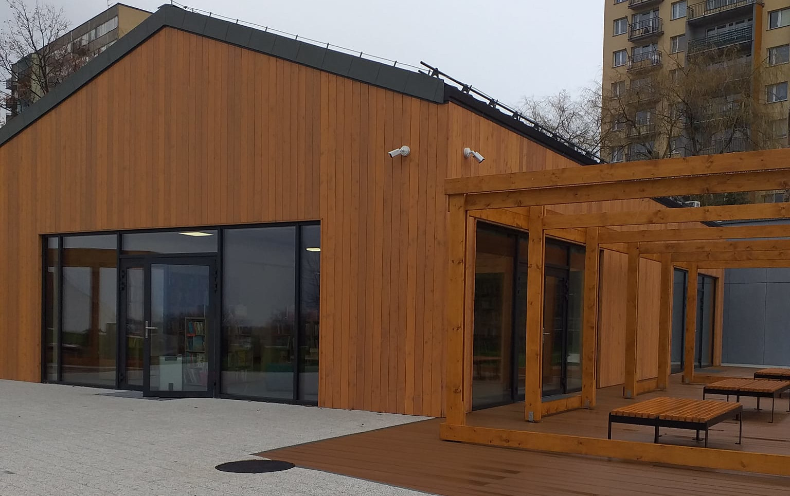 Wejście do parterowego budynku z przeszkonym wejściem i dużymi witrynami. Z prawej strony taras z ławkami