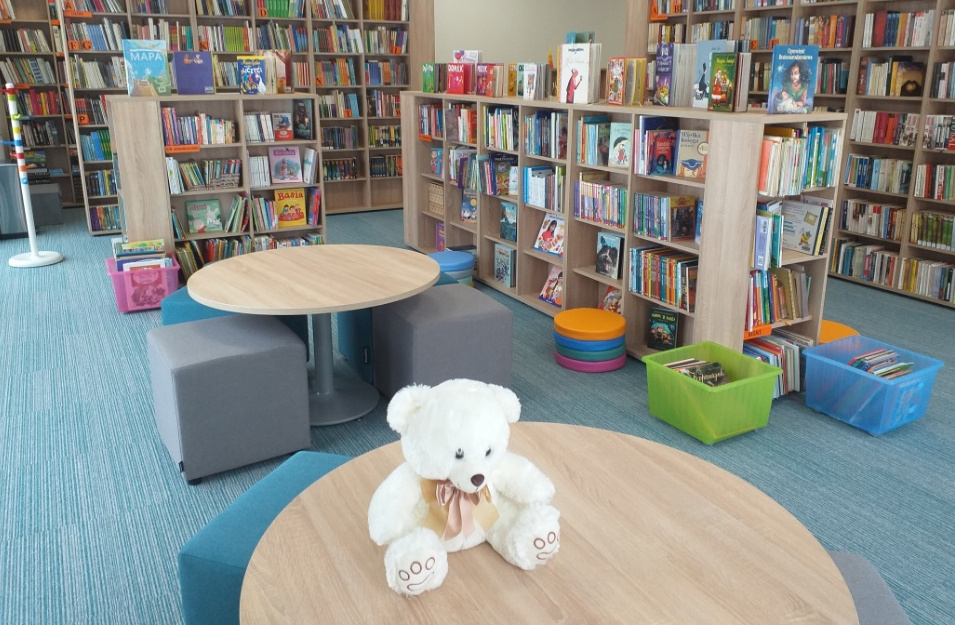 Wnętrze biblioteki. Na okrągłym stoliku biały miś, w głębi niskie regały z książkami dla dzieci