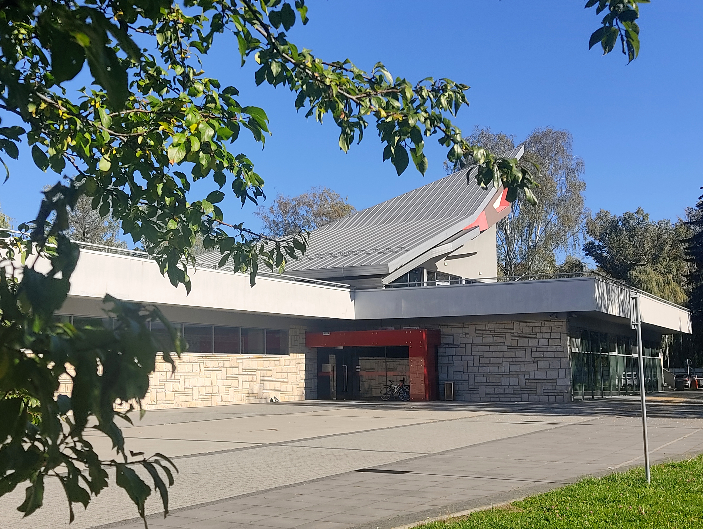 Wejście do parterowego nowoczesnego budynku. Na pierwszym planie gałęzie drzewa