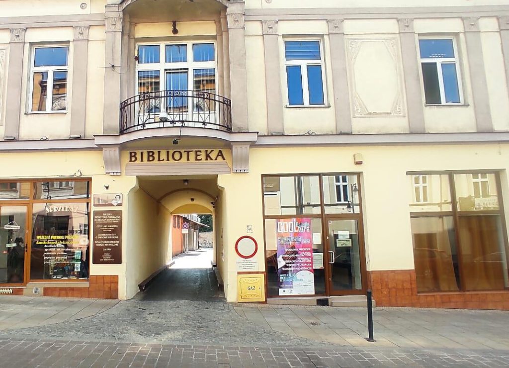 Widok na kamienicę z witrynami na parterze i półokrągłym balkonem nad bramą wjazdową Poniżej balkonu napis Biblioteka.