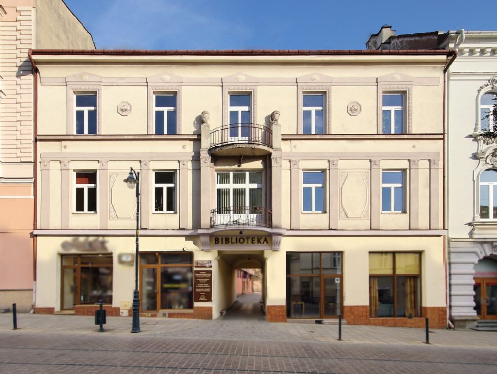 Widok na kamienicę z witrynami na parterze i półokrągłym balkonem nad bramą wjazdową Poniżej balkonu napis Biblioteka.
