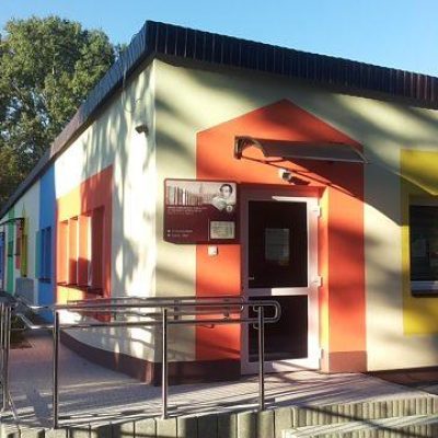 Obramowane szerokim pomarańczowym pasem wejście z logo biblioteki do niskiego parterowego budynku Z lewej i prawej strony kolorowo obramowane okna Z boku podjazd dla wózków