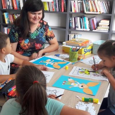 Przy stoliku czytelni bibliotekarka i trójka dzieci z kolorowankami.