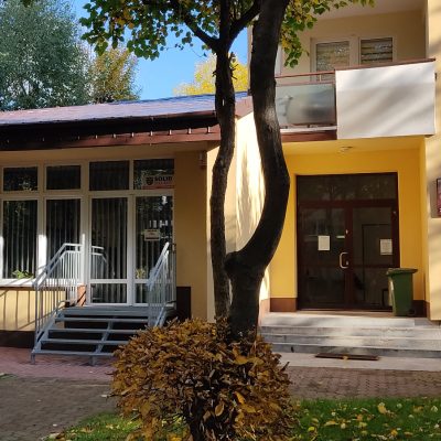 Metalowe schody do przeszklonego wejścia. Obok brama i szyld biblioteki