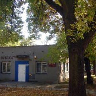 Szary parterowy budynek z dwoma oknami i płaskim dachem pod rozłożystymi starymi drzewami