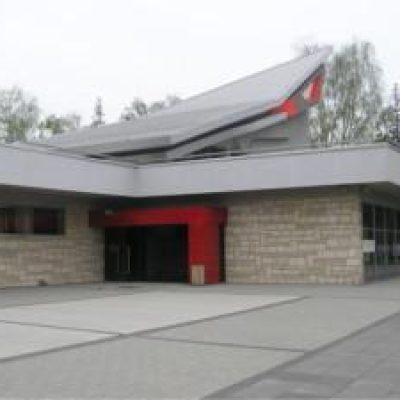 Obramowane na czerwono wejście do budynku Widoczny fragment ukośnego dachu Całość w szaro- beżowej kolorystyce