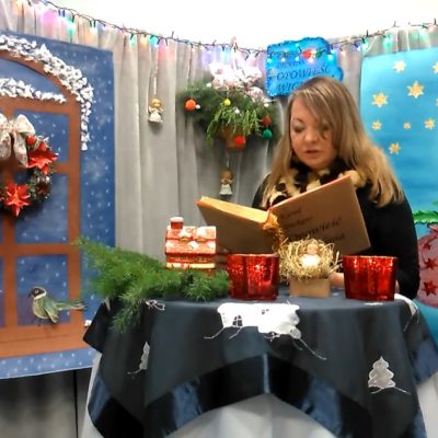 Na tle świątecznych dekoracji siedzi kobieta z długimi włosami. Trzyma otwartą książkę. Przed nią okrągły stolik nakryty ciemną serwetą z białymi świątecznymi aplikacjami. Na nim choin, żłobek i świece..